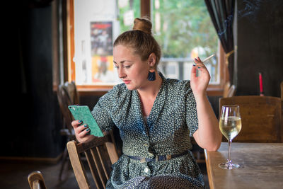 Woman smoking cigarette while using mobile phone by drink in glass on table at restaurant