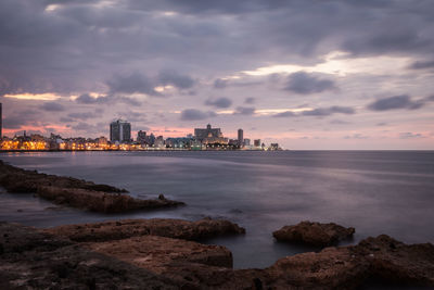 Scenic view of sea by buildings against cloudy sky at dusk in city