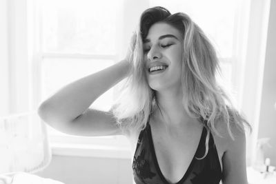 Cheerful woman wearing black bra with hand in hair at bedroom during sunny day