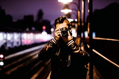 Man photographing through camera while standing at illuminated railroad station during dusk