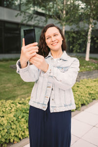Young woman in white denim jacket taking selfie on street. office building in the background