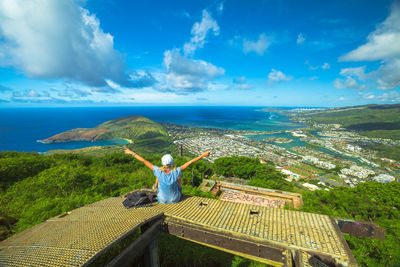 Rear view of woman with arms outstretched sitting on tower with sea in background against blue sky