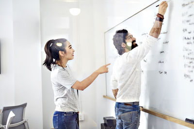 Happy businessman writing on whiteboard while businesswoman pointing in office seen through window