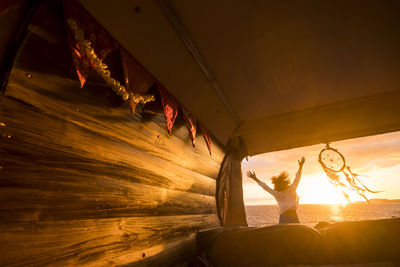 Rear view of woman with arms raised seen through vehicle at beach during sunset