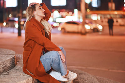 Young woman looking away while sitting on street in city at night