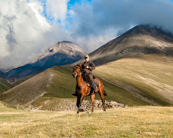 Side view of a cowgirl shepherd riding on a horse on a mountain valley against sky with clouds