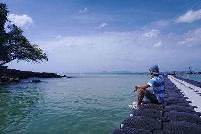 Side view of man sitting on pier over sea against cloudy sky