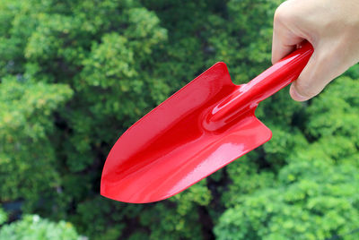 Hand holding a red shovel with blurry green foliage in the backdrop