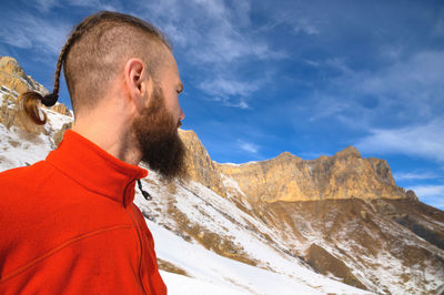 A bearded man with a stylish haircut in a red sweater stands with his back and looks into mountains 