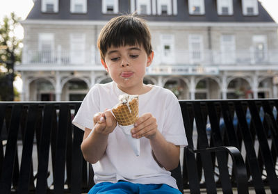 Cute boy eating ice cream cone while sitting on bench against building in city