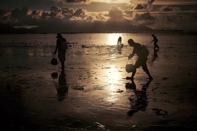 Silhouette people with metal detector on beach against sky during sunset