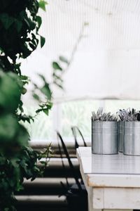 Close-up of forks and knives standing in steel jars on white table by green plants 