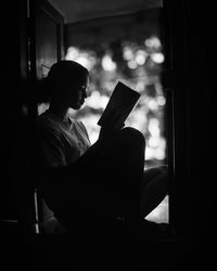 Full length of woman reading book while sitting on window