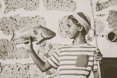 Smiling boy holding toy fish and rod against stone wall