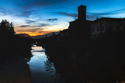 Silhouette buildings by lake against sky at sunset