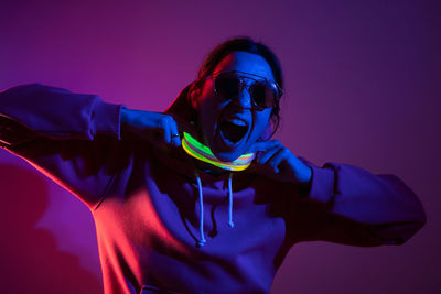 Cyberpunk woman in a hooded hoodie and sunglasses dances against a wall with neon sticks hanging 