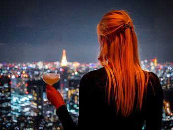 Rear view of woman having drink against illuminated cityscape at night