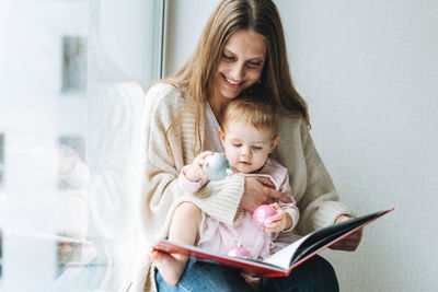 Cute little baby girl in pink dress with her mother young woman reading book in room at the home