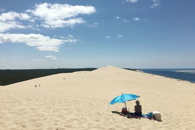 Rear view of woman sitting by umbrella at beach against sky