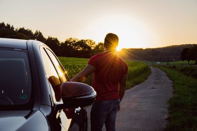 Rear view of man standing by car on road at sunset