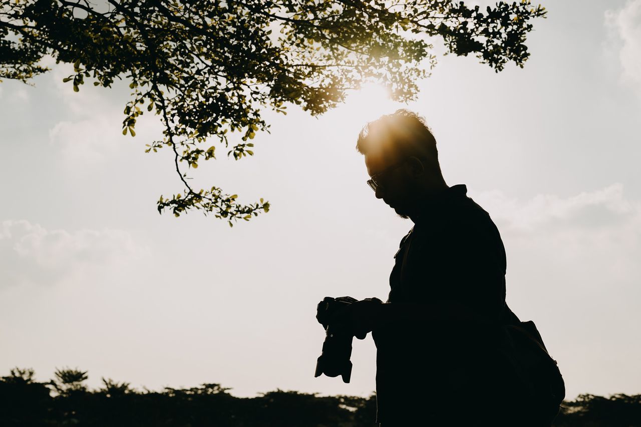 sky, silhouette, tree, plant, one person, real people, nature, lifestyles, leisure activity, sunlight, growth, men, standing, three quarter length, beauty in nature, outdoors, sunset, low angle view, day