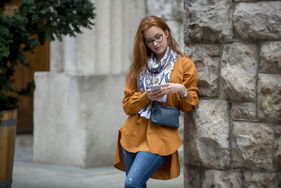 Young woman using smart phone while standing by wall