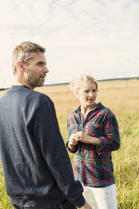 Man and woman looking away while standing on grassy field against sky