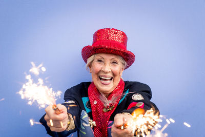 Portrait of happy senior woman wearing hat holding sparklers against blue background