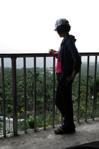 Side view of boy standing on railing against sky