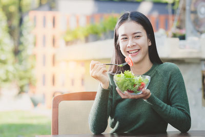 Smiling young woman holding salad bowl while sitting at table