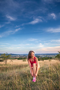 Woman tying shoelace while crouching on grass against sky