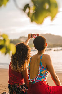 Back view of unrecognizable young ethnic women taking selfie on mobile phone while enjoying summer vacation together on sandy seashore in costa rica