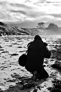 Rear view of man photographing while kneeling on sand against sky