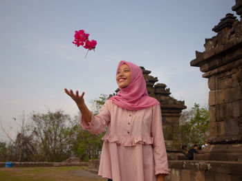 Smiling young woman playing with flowers while standing against sky