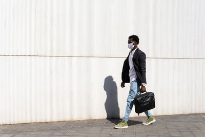 Side view of well dressed black male entrepreneur in medical mask walking along urban building on sunny day in city during coronavirus epidemic