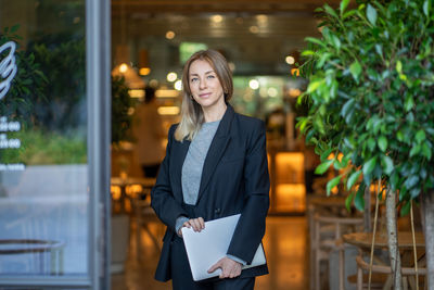 Confident entrepreneur woman stands outside contemporary cafe holds laptop, female restaurant owner
