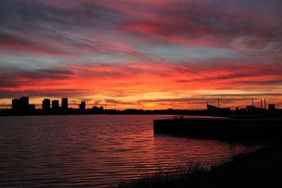 Silhouette buildings by river against sky during sunset