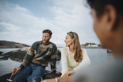 Man and woman talking with each other while sitting on rock near lake