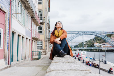 Young woman sitting on bridge over river in city