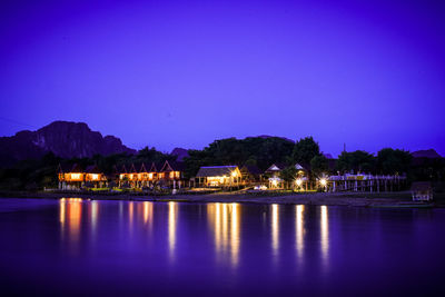 Illuminated buildings by lake against blue sky at dusk