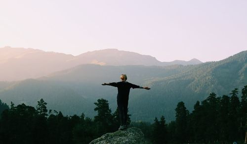 Rear view of woman with arms outstretched standing against mountains during sunset