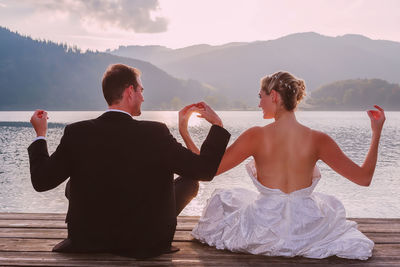 Rear view of newlywed couple sitting on pier over lake against sky during sunset