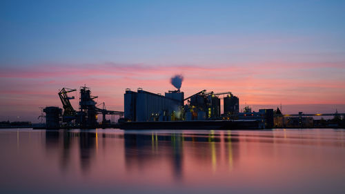 Industrial buildings by river against sky during sunset