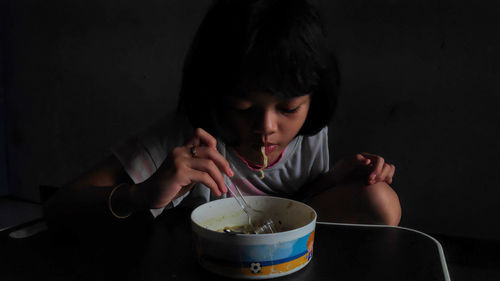 Cute asian child girl eating delicious instant noodles with fork in the dark background