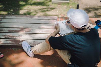 Rear view of boy drawing on book while sitting on bench outdoors