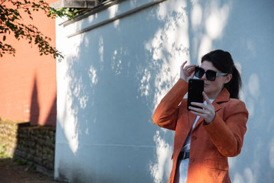 A young brunette girl in an orange jacket takes a selfie against a brick wall