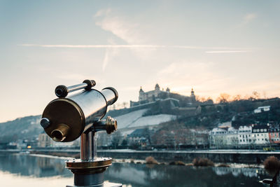 Coin-operated binoculars by river in city against sky