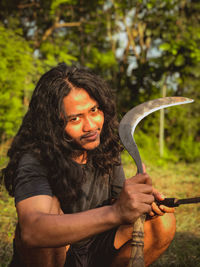 Portrait of man holding knife while crouching on land against trees