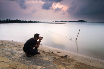 Full length of man photographing on lake against sky during sunset