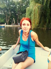 Portrait of beautiful young woman sitting by lake against trees